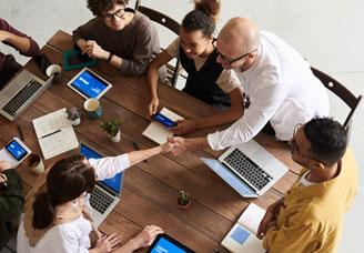 overhead photo of 6 people sitting around a table with computers & devices, shaking hands